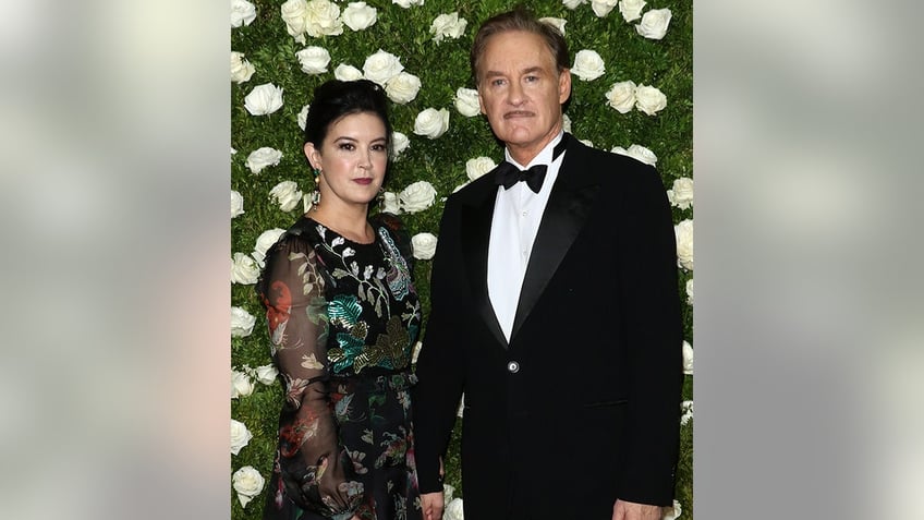 Phoebe Cates in a black patterned dress with sheer sleeves poses on the carpet with husband Kevin Kline in a classic tuxedo