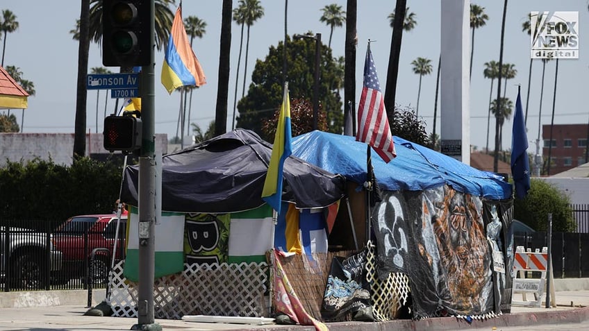 A homeless man in a clear plastic poncho stands next to a shopping cart