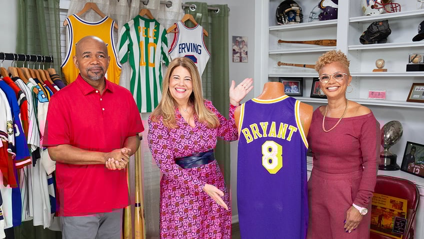 Lisa Whelchel smiling in a bright pink dress displaying a sports collection with two people.