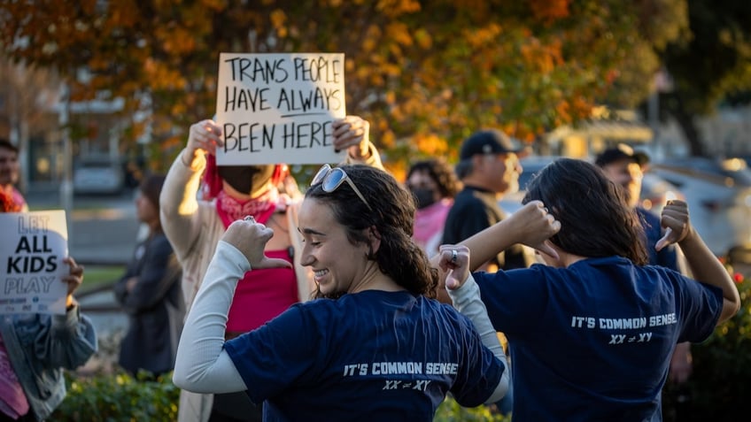 Transgender athlete supporters hold up signs at left as Tori Hitchcock, center, of the Young Women for America, and Salomay McCullough, right, show off their "Save Girls Sports" shirts.