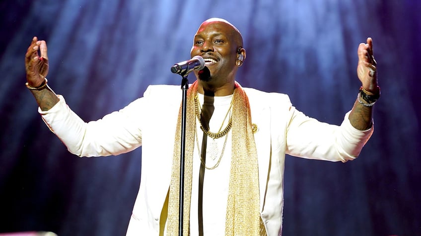 Tyrese Gibson in a white suit and gold scarf lifts his hands in the air like he's preaching on stage