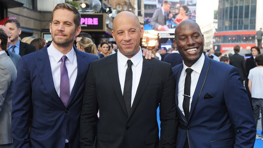 Paul Walker in a dark suit and purple tie stands next to Vin Diesel in a black suit and tie and Tyrese Gibson in a blue suit