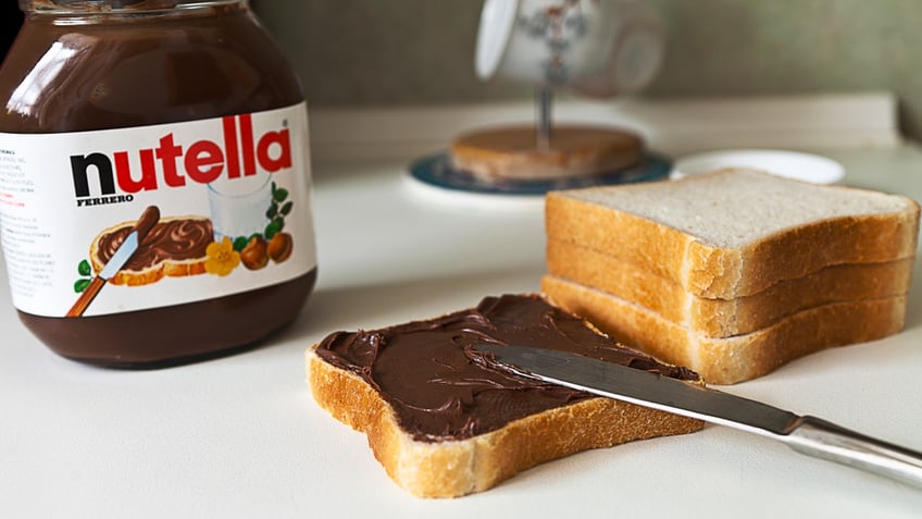 Nutella is shown on a white table next to three slices of bread and a slice of bread with a knife and the hazelnut spread on it.