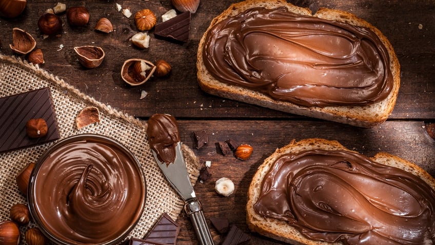 Top view of two toasts with chocolate and hazelnut spread shot on rustic wooden table. A glass bowl filled with chocolate spread and a knife are beside the toasts. Some shelled and peeled hazelnuts and chocolate pieces complete the composition.