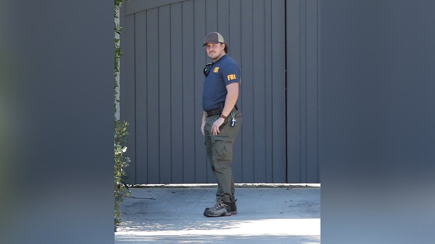 A FBI agent in a blue vest looks directly at the cameras as he stands in the driveway of Ben Affleck's home