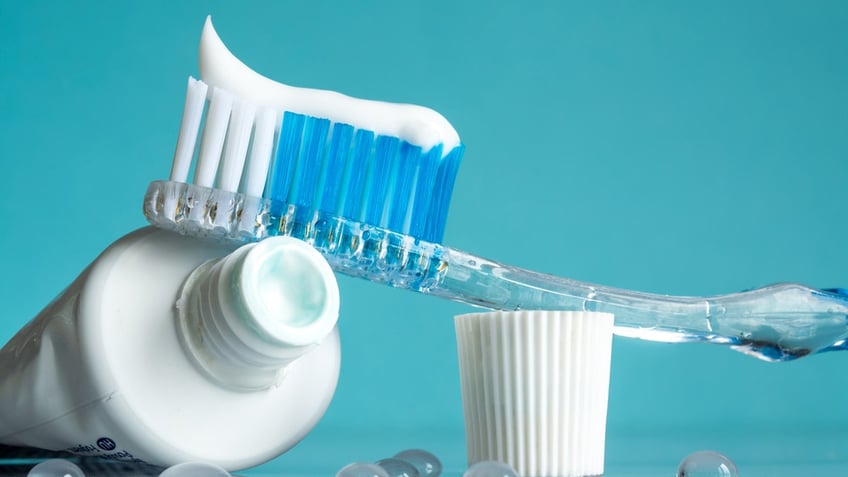 Close-up view of open toothpaste tube, cap and tooth brush in front of a blue backdrop.