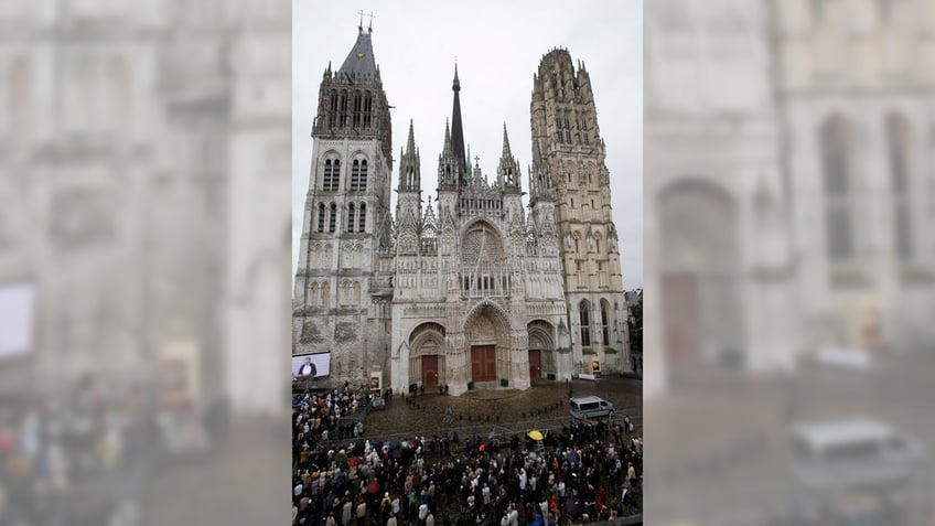 A crowd stands outside of the the Rouen cathedral on an overcast day.