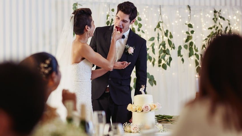 A happy couple taste the wedding cake at their reception in front of guests.