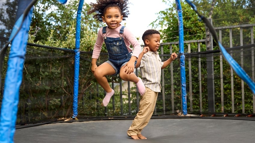 Boy And Girl Having Fun Playing In Garden Bouncing On Trampoline