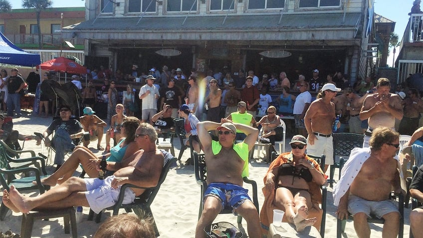 People sit and stand while mingling on the sand at the beach outside a bar.