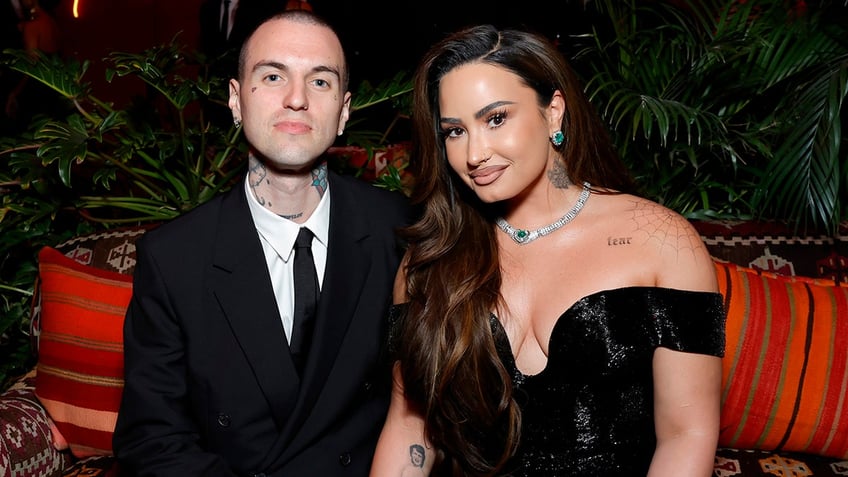 Jordan Lutes in a black suit and tie soft smiles with Demi Lovato in a plunging off the shoulder black gown at the Vanity Fair Oscar Party