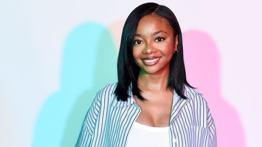 Skai Jackson in a jean jacket and white shirt smiles for the camera with a pink and blue shadow of her behind