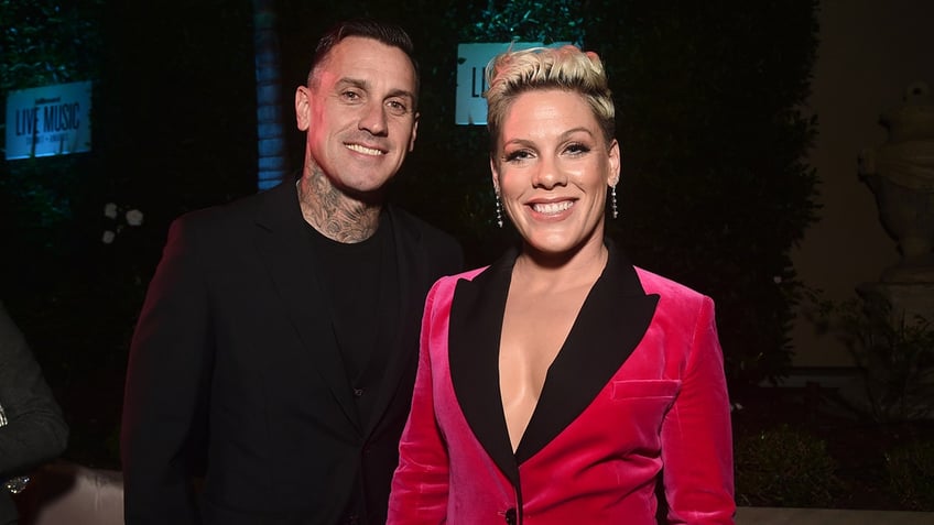 Carey Hart smiles for a photo in black with wife Pink in a pink velvet suit jacket with black lapels