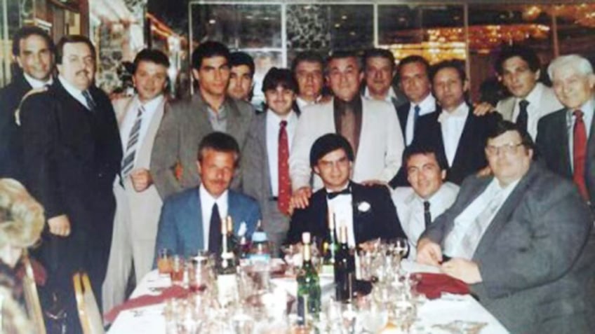 Michael Franzese in a suit and glasses being surrounded by men at a dinner table.