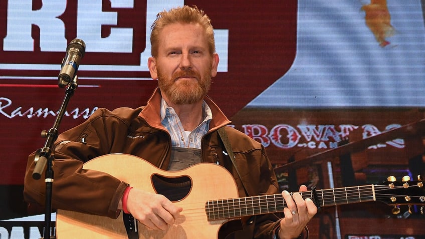 Rory Feek in a brown jacket looks up on stage as he plays the guitar