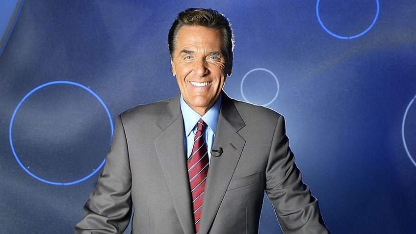 Chuck Woolery in a grey suit, blue shirt and red striped tie smiles for a photo