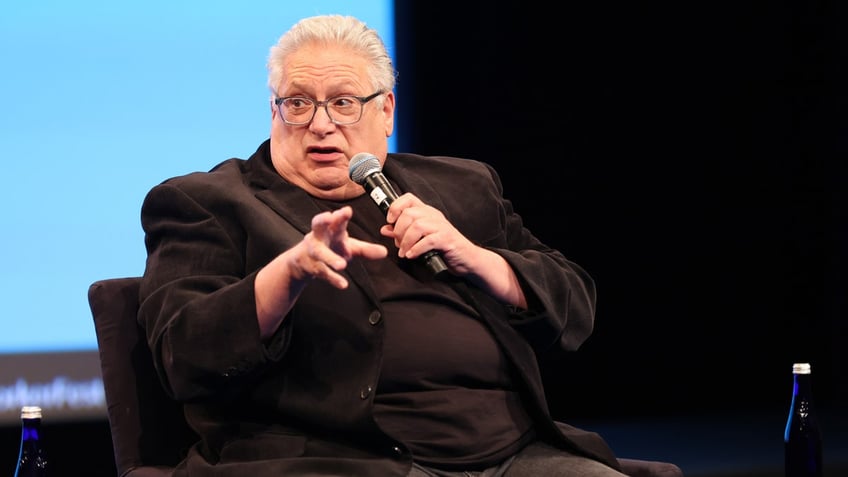 Harvey Fierstein in a black shirt and suit speaks on stage in a chair