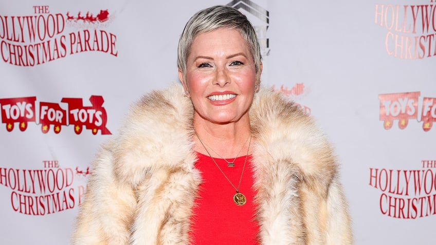 Nicole Eggert in a bright red shirt and fuzzy fur-like coat smiles on the carpet