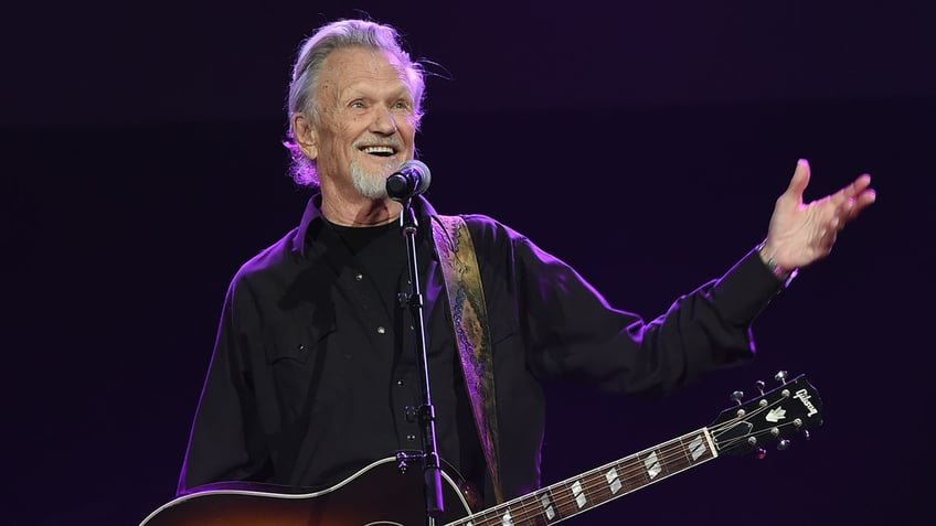 Kris Kirstofferson smiles in a black shirt on stage with his guitar on his shoulder