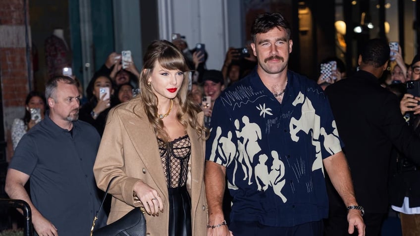 Taylor Swift in a brown jacket and black purse holds Travis Kelce's hand in a navy blue shirt with shapes of figures/animals drawn on it