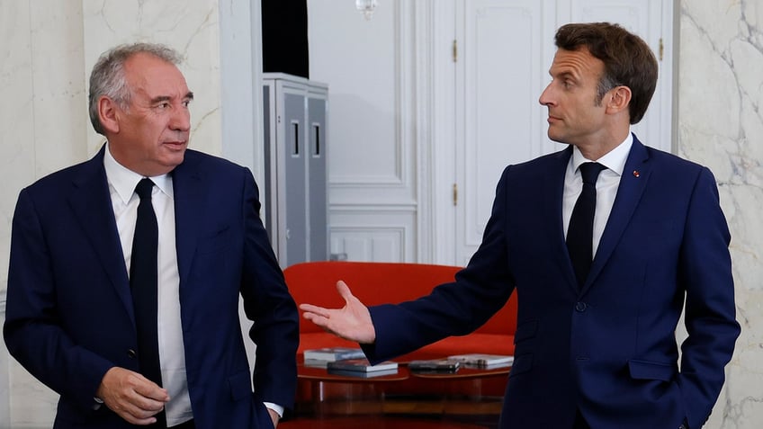 French President Emmanuel Macron holds his hand out to his centrist ally, Francois Bayrou. Both men wear blue suits, white shirts, and black ties.