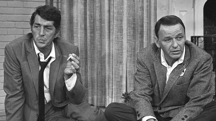 Dean Martin holding a cigarette and sitting next to Frank Sinatra as they both look concerned.