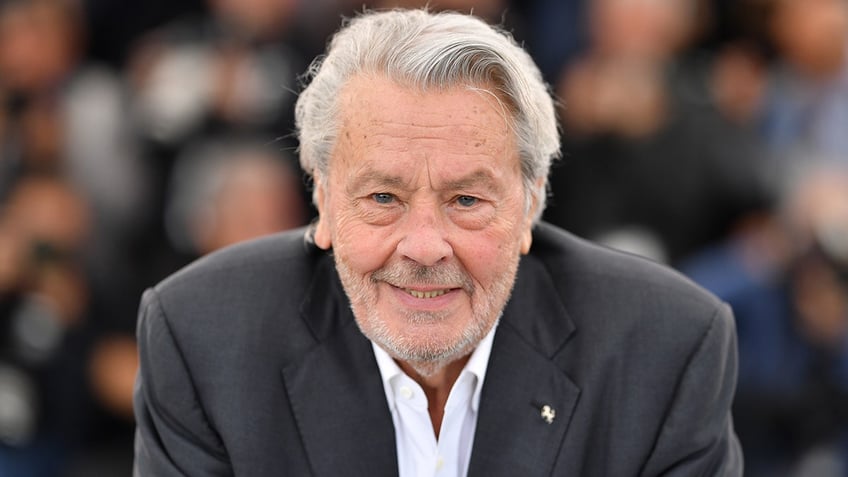 Alain Delon smiles in a black suit and white shirt in Cannes