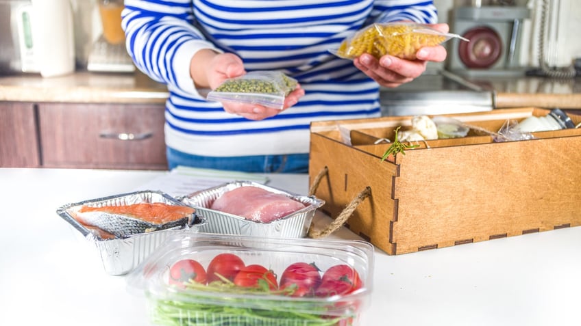 A woman's hands holds a bag of pasta and greens while taking apart a delivery box with fresh ingredients.