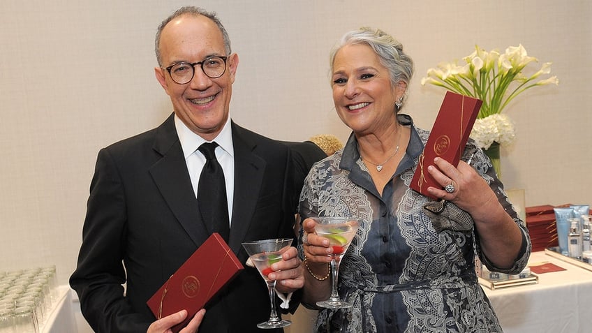 David Crane in a black suit and Marta Kauffman in a printed dress, both holding drinks, smile for the camera