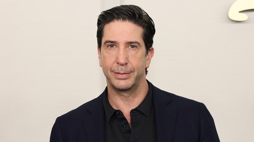 David Schwimmer in a black suit and shirt looks serious on the carpet