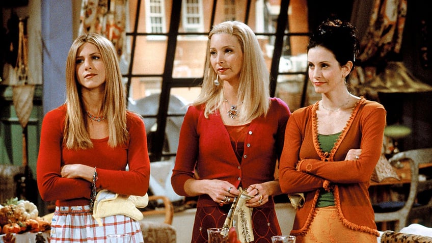 Jennifer Aniston and Lisa Kudrow in red as Rachel and Phoebe on Friends standing next to Courteney Cox as Monica in orange, all looking unamused