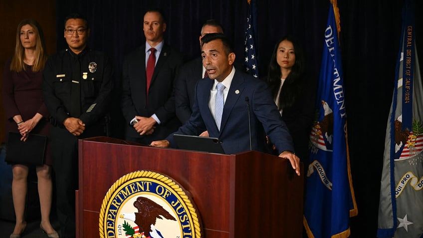 US Attorney for the Central District of California Martin Estrada in a blue suit speaks behind a podium with many people standing behind him.