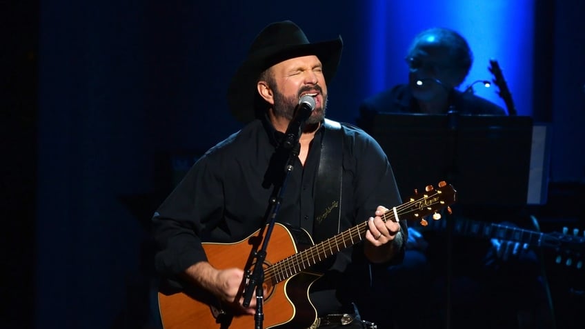 Garth Brooks performs on stage with a guitar in all black outfit