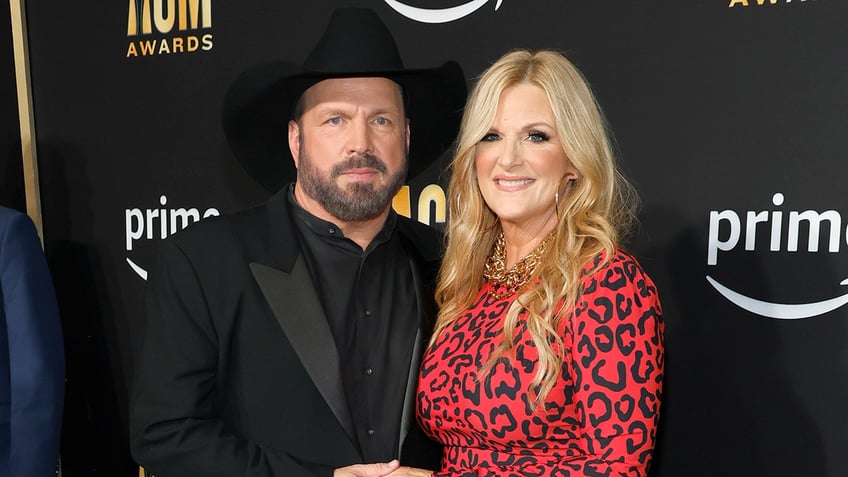 Garth Brooks in a black suit and cowboy hat poses with wife Trisha Yearword in a red cheetah print dress on the carpet