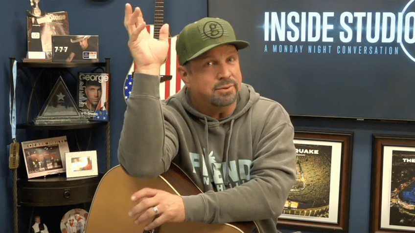 Garth Brooks in a sweatshirt and green baseball cap puts his hand in the air as he rest the other over his guitar