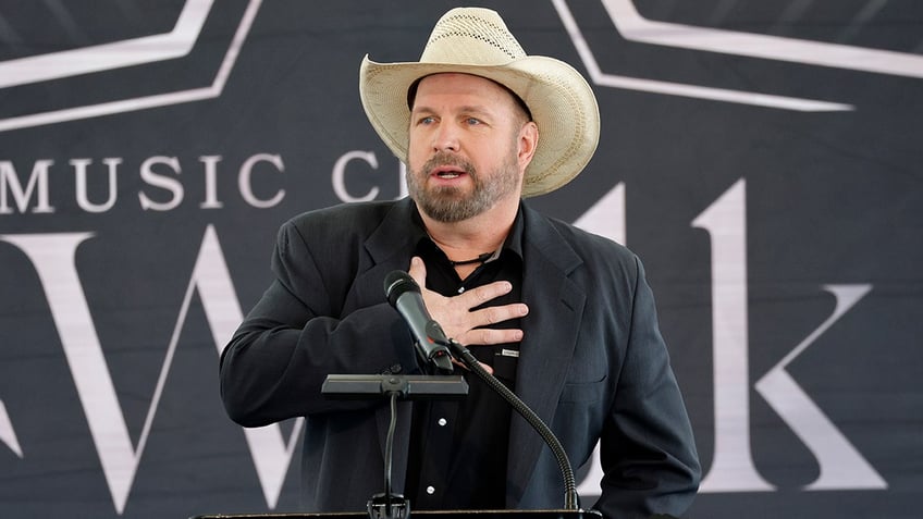 Garth Brooks puts his hands to his chest behind a podium wearing a black suit and tan cowboy hat