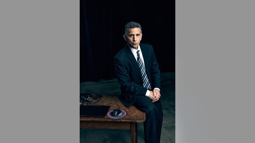 George Olivo in a dark suit sitting on a desk.