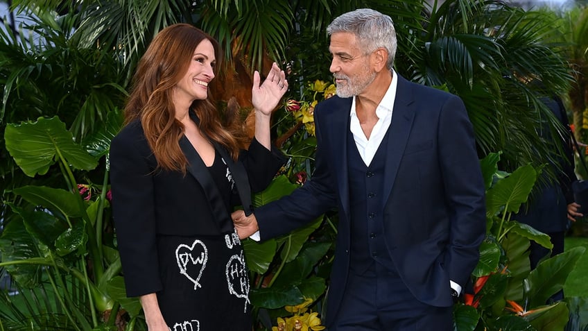 Julia Roberts in a black blazer with hears smiles as she greets George Clooney on the carpet in a blue suit