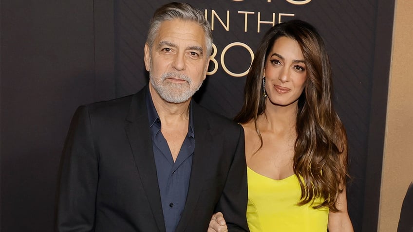 George Clooney in a black blazer and blue button down stands next to wife Amal in a yellow gown