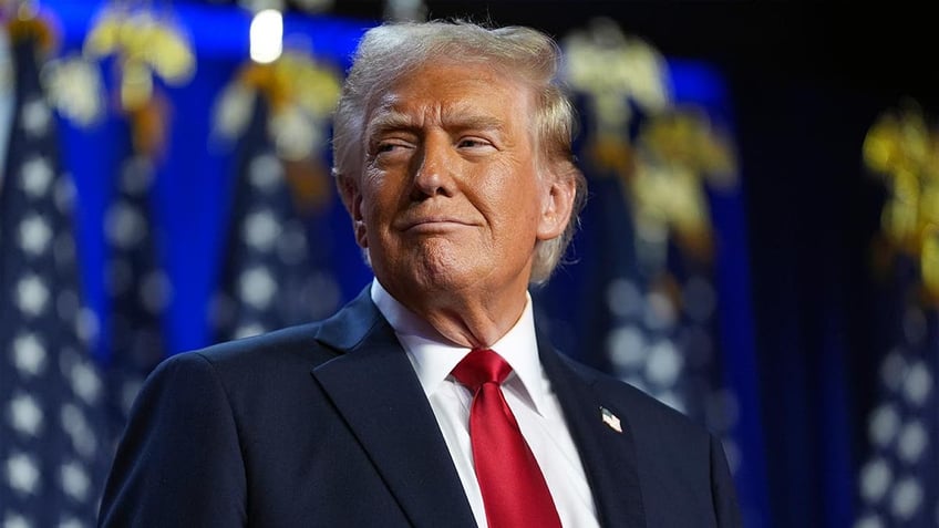 Donald Trump smiling in closeup shot, flags behind him