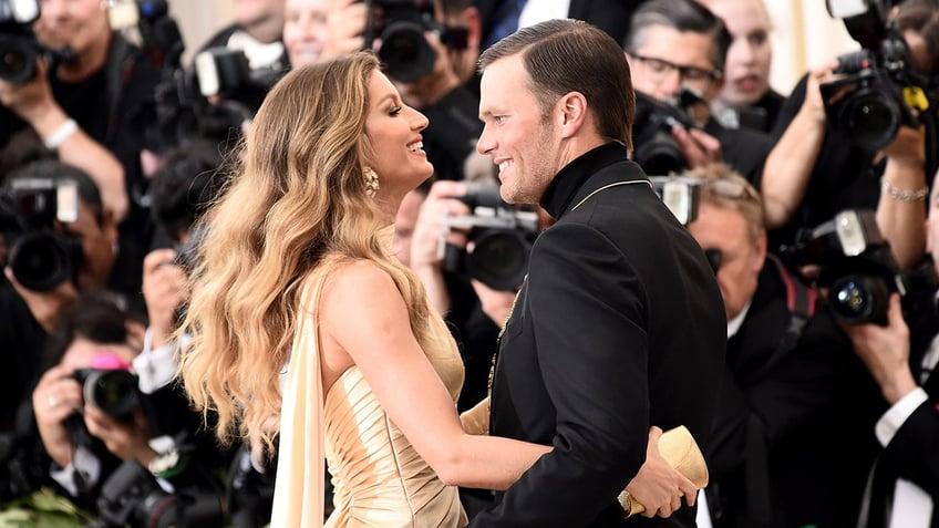 Gisele Bündchen in a gold dress looks lovingly at Tom Brady in a black suit