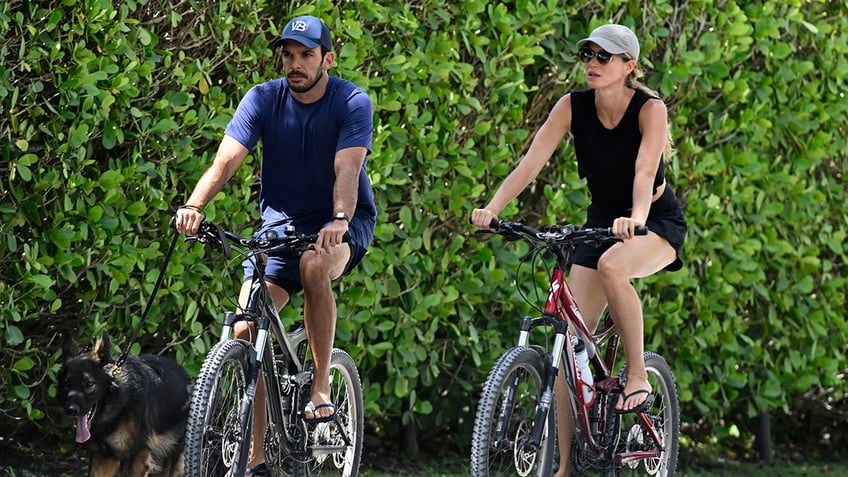 Joaquim Valente in a blue shirt and hat rides his bike alongside Gisele Bündchen in a black outfit on her bike