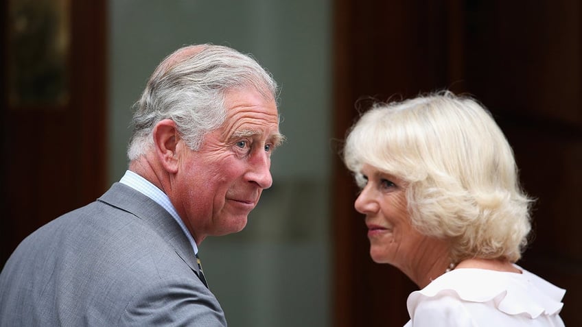 Queen Camilla wearing a white dress looking at King Charles wearing a grey suit.