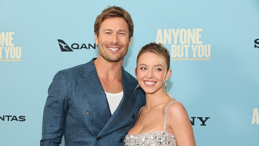 Glen Powell in a blue blazer smiles widely on the carpet with Sydney6 Sweeney in a beaded light blue/silver dress