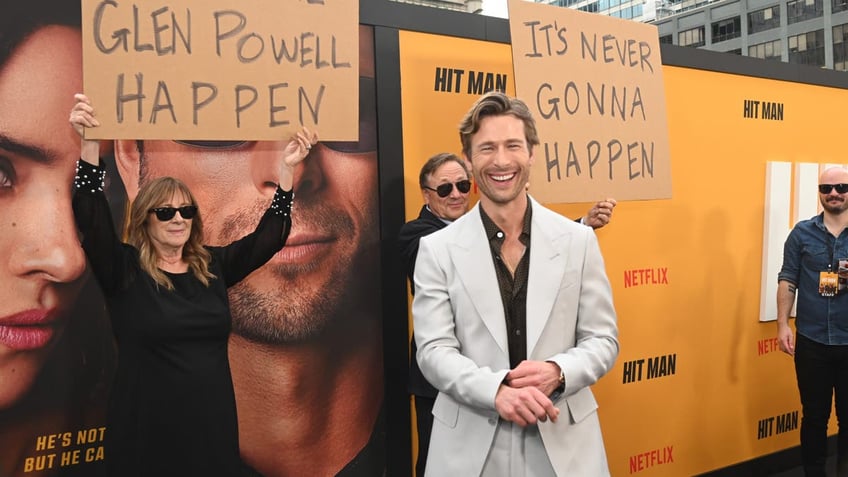 glen powell on red carpet smiling in front of parents holding signs