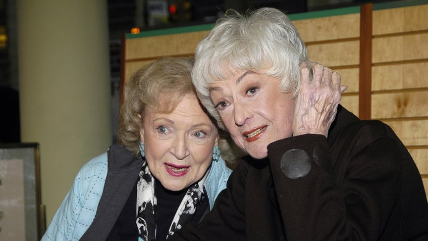 Betty White in a blue jacket leans against Bea Arthur in a brown jacket