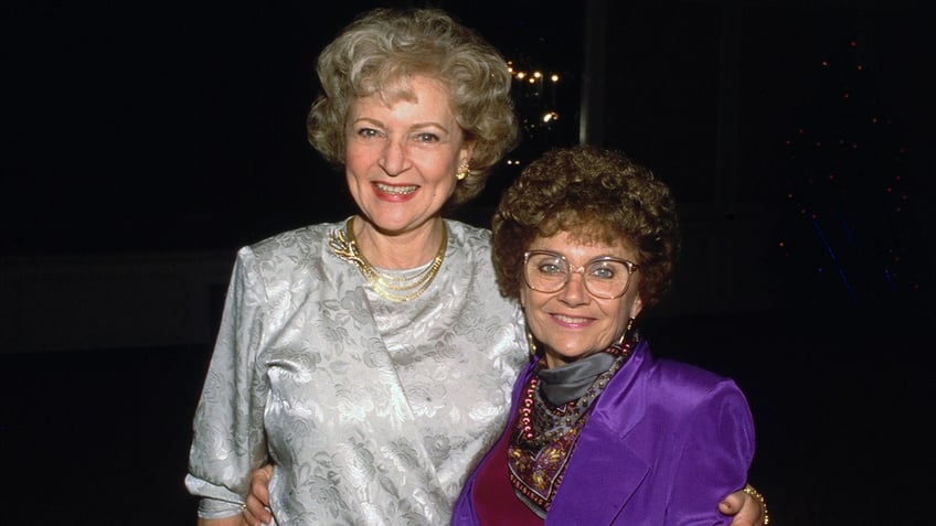 Betty White in silver floral dress smiles next to Estelle Getty in a purple dress and patterned scarf
