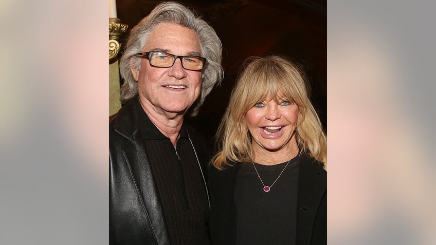 Kurt Russell in a sweater and black leather jackets smiles next to a happy Goldie Hawn in a black sweater and jacket and pink-stoned necklace