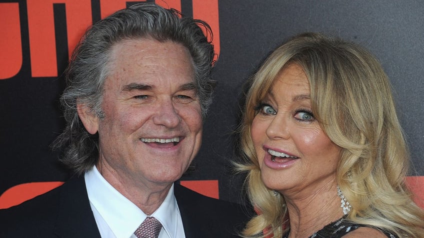 Kurt Russell in a black suit smiles next to Goldie Hawn who makes a surprised/silly face on the carpet