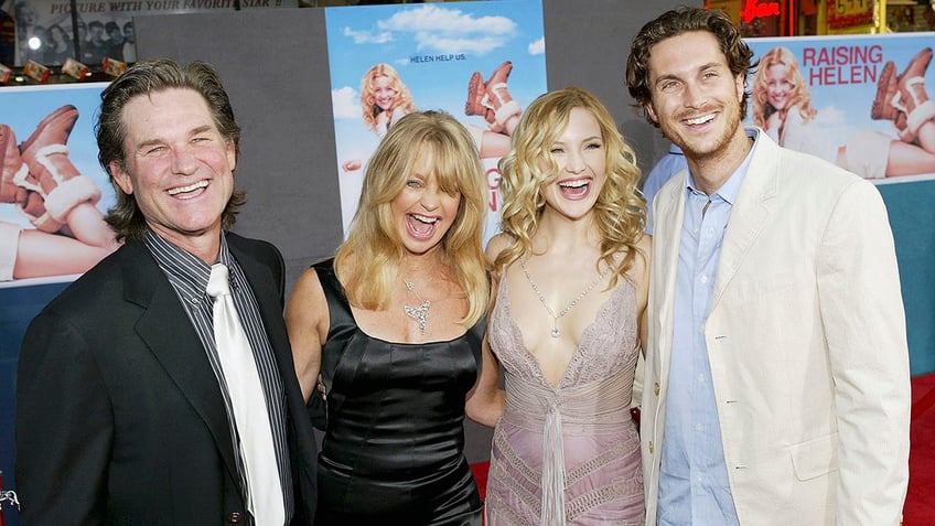 Kurt Russell in a black suit and black and white striped shirt laughs with Goldie Hawn ina. black dress, Kate Hudson in a light pink lacy dress and Oliver Hudson in a cream jacket on the carpet